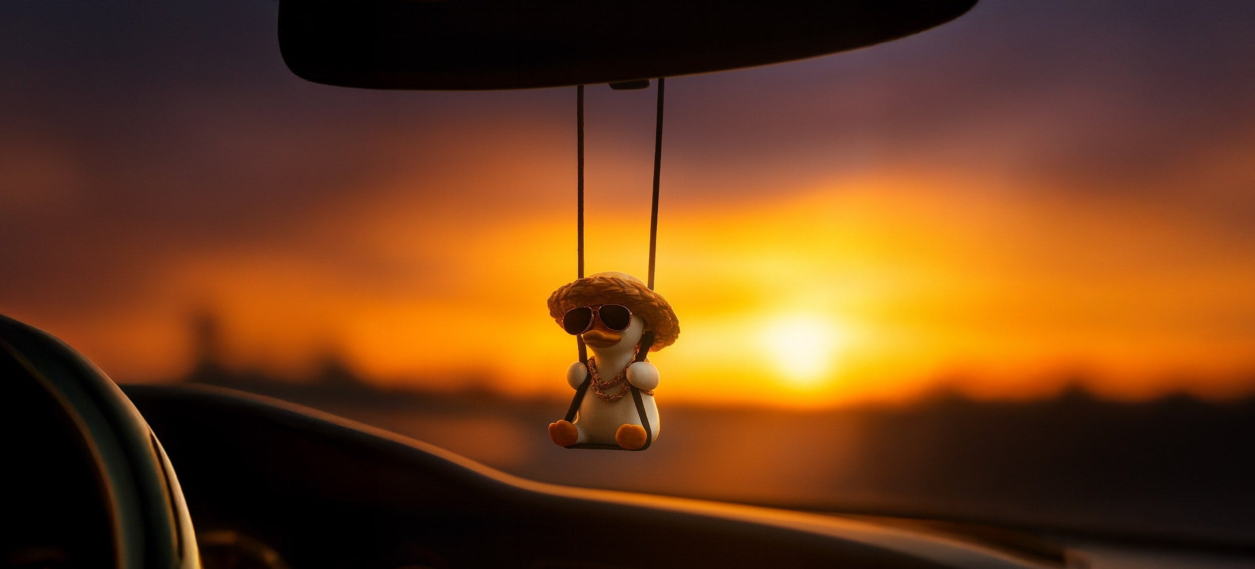 Car interior with a small figurine hanging from the rearview mirror against a sunset sky.